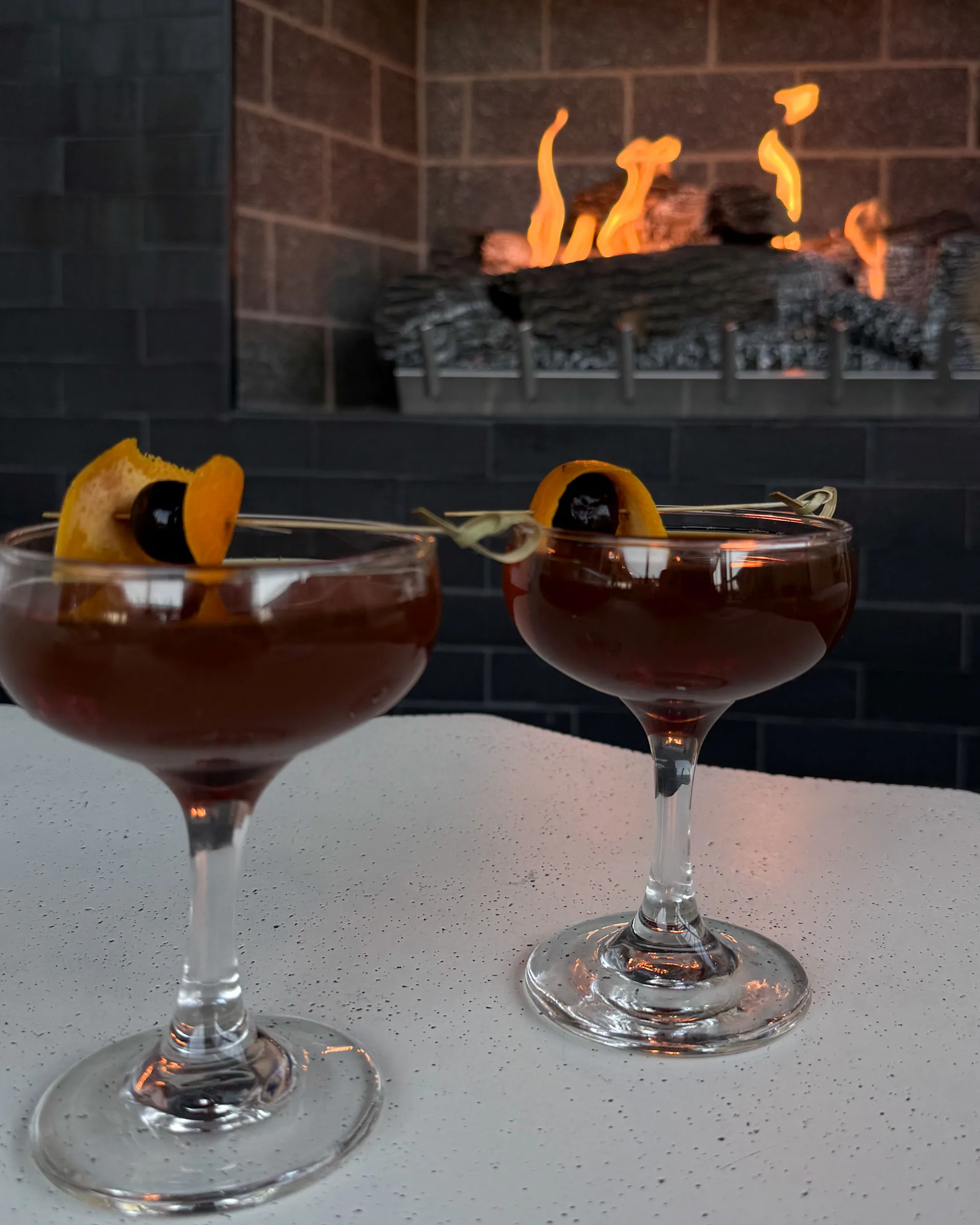 Two espresso martinis with an orange garnish on a table in front of a lit fireplace.