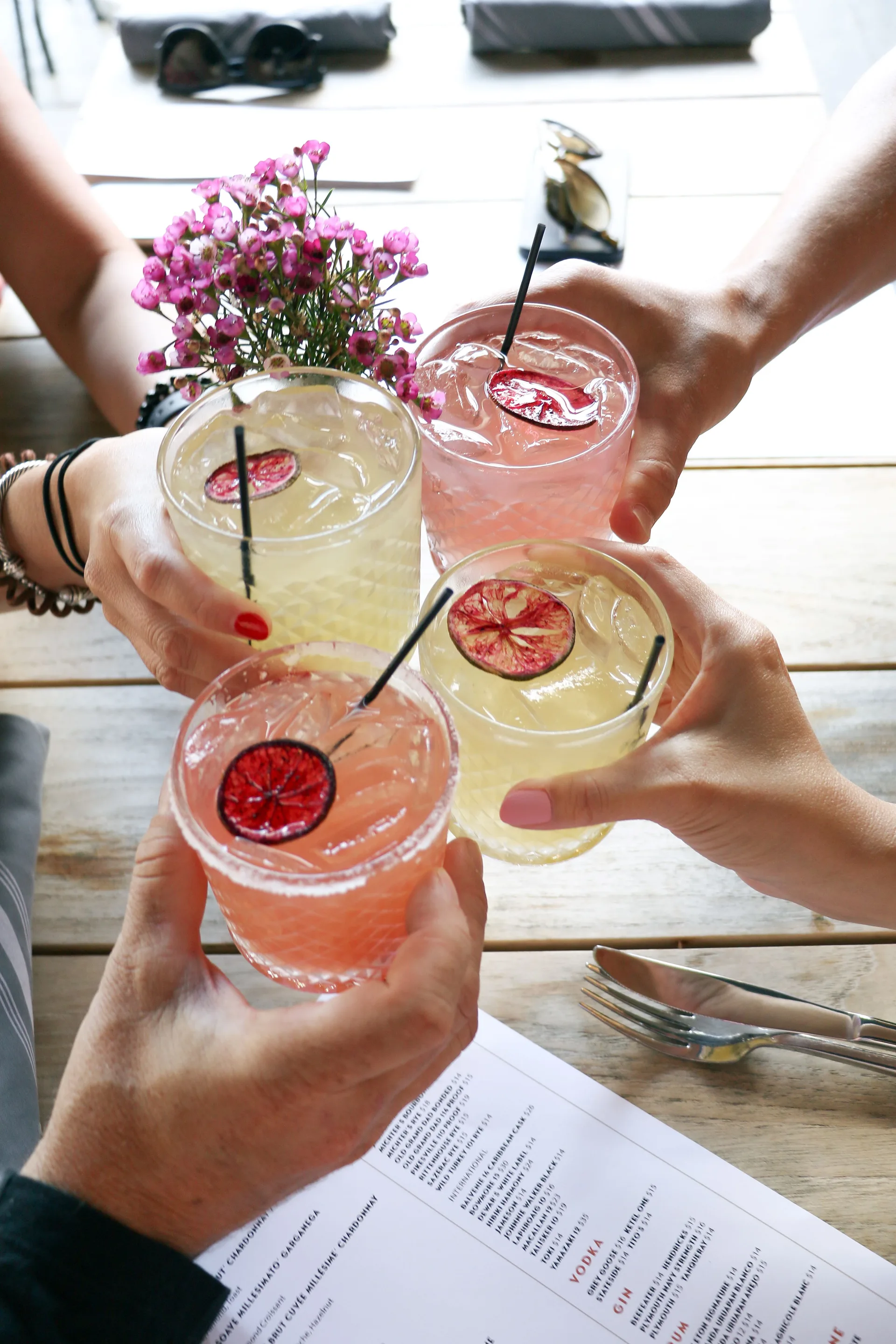 4 hands holding cocktails that are either yellow or pink, with a purple flower centerpiece in the middle.