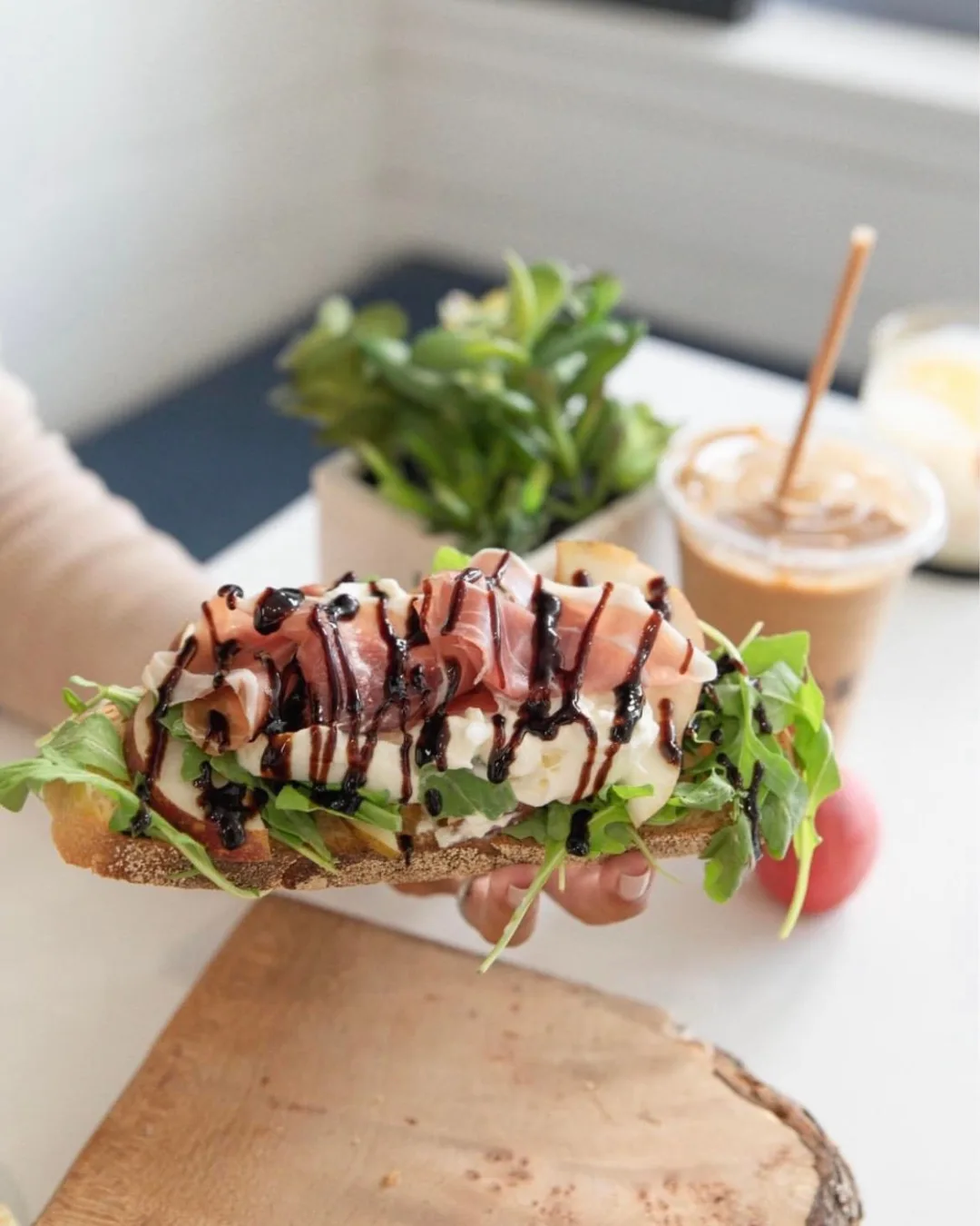 A hand holding an avocado toast next to an iced coffee.
